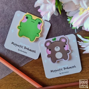 以下が含まれることがあります： 緑色のカエルと茶色のクマの2つの手作り磁石ブックマーク。両方のブックマークはピンクの頬をしていて、白のドットがついた薄い紫色の背景になっています。ブックマークのテキストには「Handmade Magnetic Bookmark by RoroLulu」と書いてあります。