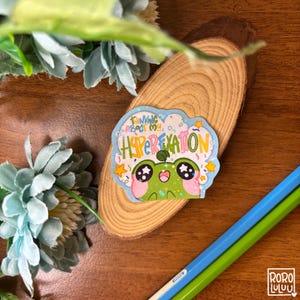 May include: A vibrant sticker with a cartoon frog character and the words "Thinking About My Hyperexcitation". The frog is green with pink cheeks, set against a light blue cloud shape with stars and sprinkles. The sticker is on a wooden slice.