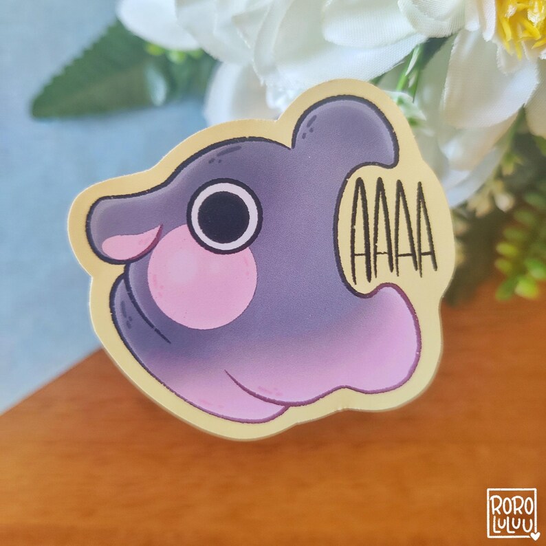 May include: A purple and pink cartoon fish with a large eye and a wide open mouth. The fish is yelling "AAAA" in black text. The sticker is on a wooden surface.