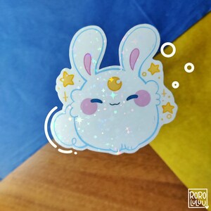 May include: A holographic sticker featuring a round, white, fluffy rabbit character with long ears, pink cheeks, and a yellow crescent moon on its forehead. The design includes small yellow stars and white circular accents, with the text RORO LULUU in the corner.