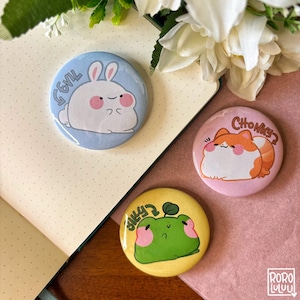 Peut inclure: Trois épingles rondes avec des animaux de dessin animé. Une épingle bleue avec un lapin blanc avec le texte "GEVIL". Une épingle rose avec un chat orange avec le texte "CHONKY?". Une épingle jaune avec une grenouille verte avec le texte "SILLY?".
