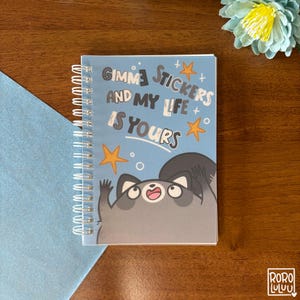 以下が含まれることがあります： 「Gimme Stickers and My Life is Yours」というテキストとアライグマのイラストが描かれた青いスパイラルノートです。