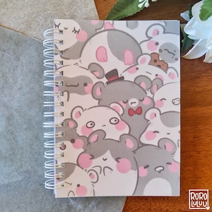 以下が含まれることがあります： ピンクの頬をしたかわいい漫画の動物のパターンが表紙に描かれた白いスパイラルバインダーノート。動物はほとんどがグレーと白で、黒い目とピンクの鼻をしています。表紙には、動物の1つに小さな黒い蝶ネクタイも付いています。