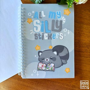 以下が含まれることがあります： 「ALL my SILLY stickers」の文字と、ステッカーブックを持つアライグマのイラストが描かれた、水色の表紙のリングノート。星と円のグラフィックが表紙を飾っています。