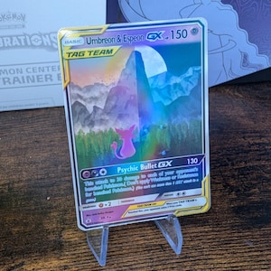 Könnte beinhalten: Eine Pokemon-Sammelkarte mit Umbreon & Espeon GX, einer Tag-Team-Karte mit holografischem Hintergrund. Die Karte hat ein lila und blaues Farbschema und zeigt eine Bergkette mit einem Vollmond im Hintergrund. Die Attacke der Karte ist Psychischer Geschoss GX, die jedem Pokémon auf der Bank deines Gegners 130 Schaden zufügt.