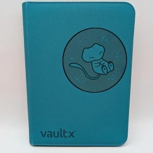 May include: Teal Vault X binder with a circular graphic of a cartoon creature. The binder has a stitched border and the Vault X logo at the bottom. The binder is closed and appears to be new.