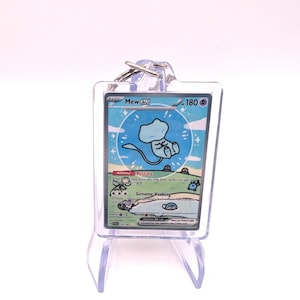 May include: A clear acrylic keychain featuring a Pokémon trading card with a blue Mew character. The card has the text "Mew ex" and "180" on it. The keychain is displayed on a clear plastic stand.