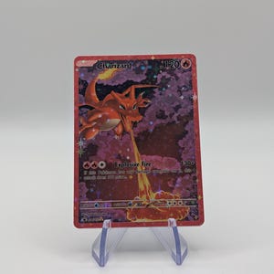 May include: A Charizard Pokémon trading card, featuring a red and orange dragon-like creature breathing fire. The card has a holographic background and text that reads "Explosive Fire". The card is displayed on a clear plastic stand.