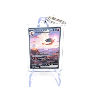 May include: A clear acrylic keyring displaying a Charizard EX trading card. The card shows a colourful illustration of a Charizard flying over a volcanic landscape, with the text "Charizard EX" and attack details.