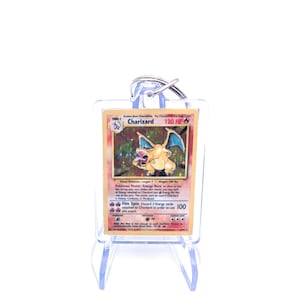Peut inclure: Un porte-clés avec une carte Pokémon Charizard. La carte présente une illustration de Charizard avec le texte "Charizard" et "120 HP". Le porte-clés est enfermé dans un support de protection transparent et attaché à un porte-clés argenté.