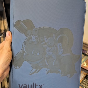 May include: A blue rectangular binder with a textured surface featuring a dark graphic print of the Pokemon characters Blastoise, Squirtle, and Raichu. The word vaultx is printed in dark letters at the bottom center of the front cover.