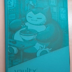 Peut inclure: Classeur Vault X turquoise avec une illustration de Snorlax mangeant des ramen. L'œuvre d'art est dans une teinte turquoise, avec du texte japonais et une scène de restaurant. Le classeur a une fermeture éclair et le logo Vault X.