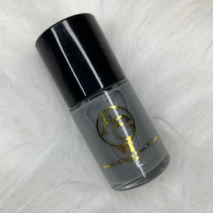 May include: A bottle of Avant Garde nail polish in a dark gray color with a black cap. The bottle has a gold logo and text. The nail polish bottle is on a white, fluffy surface.