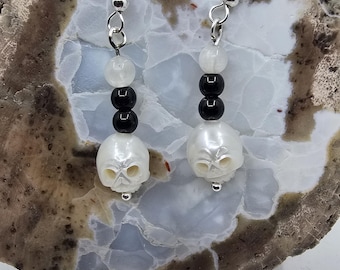 May include: A pair of dangle earrings featuring a skull design. Each earring has a silver hook, a series of black and white beads, and a white skull pendant. The earrings are displayed on a light-colored, textured surface.