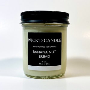 May include: A glass jar candle with a black lid and label. The label reads "WICK'D CANDLE", "HAND POURED SOY CANDLE", "BANANA NUT BREAD", "8 Oz.", and "Made in Ohio".