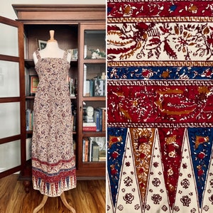 May include: A long, white maxi dress with a floral print in shades of red, blue, and brown. The dress has a square neckline and thin straps. The hemline features a decorative border with a geometric pattern.