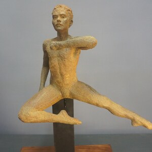 May include: A bronze sculpture of a person in a dynamic pose, with one leg extended and arms outstretched. The figure has a textured surface and is mounted on a dark metal support.