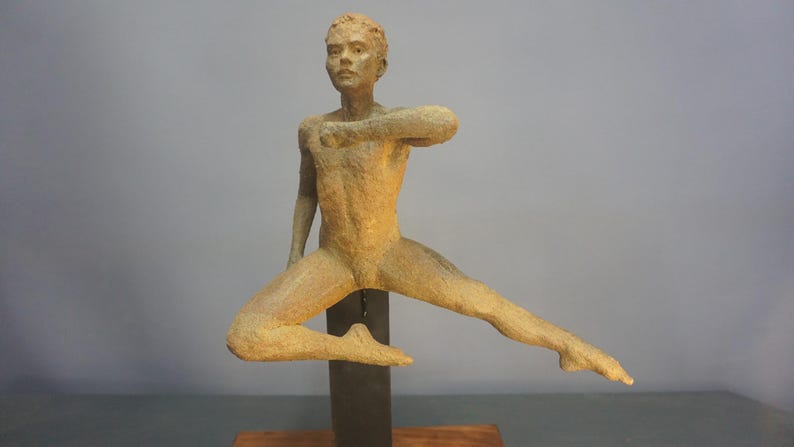May include: A bronze sculpture of a person in a dynamic pose, with one leg extended and arms outstretched. The figure has a textured surface and is mounted on a dark metal support.