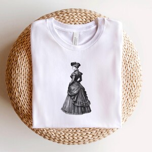 Pu&ograve; includere: T-shirt bianco con un'illustrazione in bianco e nero di una donna in abito vittoriano. La grafica &egrave; centrata. La maglietta &egrave; esposta su una superficie rotonda, intrecciata, marrone chiaro.