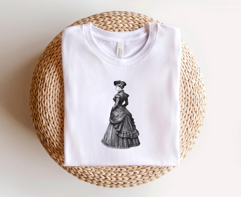 Pu&ograve; includere: T-shirt bianco con un'illustrazione in bianco e nero di una donna in abito vittoriano. La grafica &egrave; centrata. La maglietta &egrave; esposta su una superficie rotonda, intrecciata, marrone chiaro.