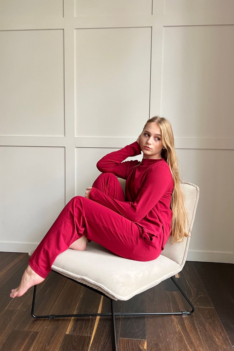 Pu&ograve; includere: Un completo rosso composto da top a maniche lunghe e pantaloni abbinati. L'outfit &egrave; in tinta unita e sembra essere realizzato in un materiale morbido. La persona &egrave; seduta su una sedia color crema con struttura in metallo nero.