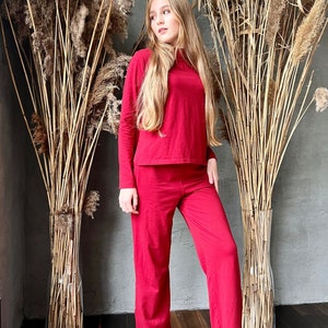 Pu&ograve; includere: Un completo rosso composto da un top a maniche lunghe e pantaloni a gamba larga abbinati. L'outfit &egrave; realizzato in un materiale morbido. La modella &egrave; in piedi davanti a piante alte e secche in un vaso. Lo sfondo &egrave; un muro grigio e un pavimento in legno scuro.