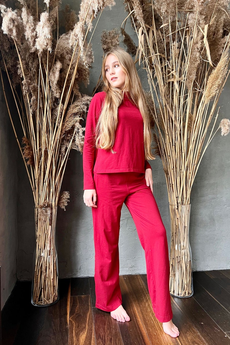 Pu&ograve; includere: Un completo rosso composto da un top a maniche lunghe e pantaloni a gamba larga abbinati. L'outfit &egrave; realizzato in un materiale morbido. La modella &egrave; in piedi davanti a piante alte e secche in un vaso. Lo sfondo &egrave; un muro grigio e un pavimento in legno scuro.