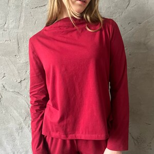 Pu&ograve; includere: Un completo pigiama bordeaux a due pezzi. La maglia a maniche lunghe ha uno scollo tondo e i pantaloni sono in tinta. L'outfit &egrave; realizzato in un materiale morbido. La modella indossa il completo davanti a un muro grigio strutturato.