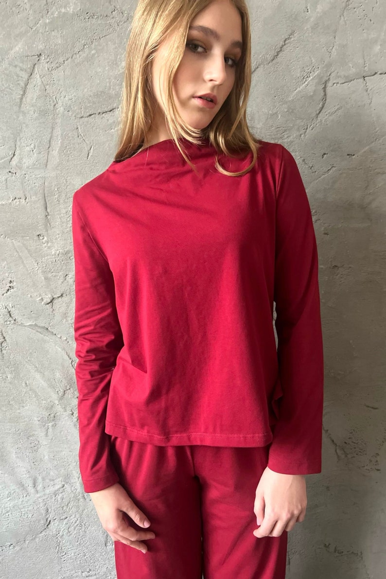 Pu&ograve; includere: Un completo pigiama bordeaux a due pezzi. La maglia a maniche lunghe ha uno scollo tondo e i pantaloni sono in tinta. L'outfit &egrave; realizzato in un materiale morbido. La modella indossa il completo davanti a un muro grigio strutturato.