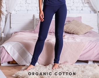 Può includere: Una persona che indossa leggings blu scuro in piedi davanti a un letto con una coperta rosa e bianca. Il testo "ORGANIC COTTON" è stampato sul pavimento davanti alla persona.