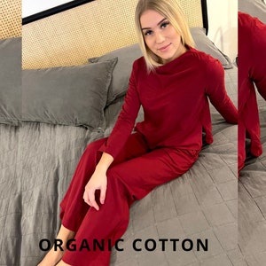 Pu&ograve; includere: Un completo pigiama rosso in cotone biologico, composto da un top a maniche lunghe e pantaloni a gamba larga. Il testo "ORGANIC COTTON" &egrave; visualizzato nella parte inferiore dell'immagine. Il completo &egrave; mostrato su un letto con biancheria da letto grigia.