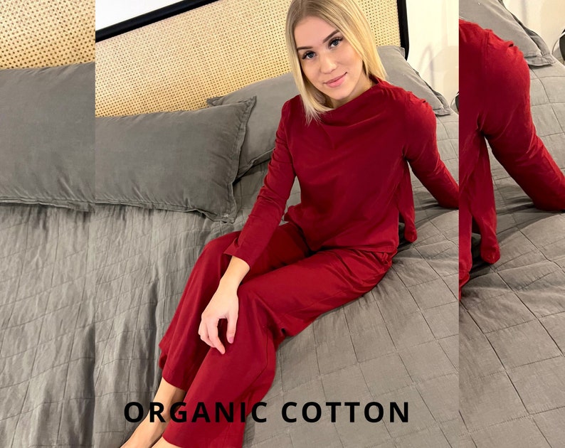 Pu&ograve; includere: Un completo pigiama rosso in cotone biologico, composto da un top a maniche lunghe e pantaloni a gamba larga. Il testo "ORGANIC COTTON" &egrave; visualizzato nella parte inferiore dell'immagine. Il completo &egrave; mostrato su un letto con biancheria da letto grigia.