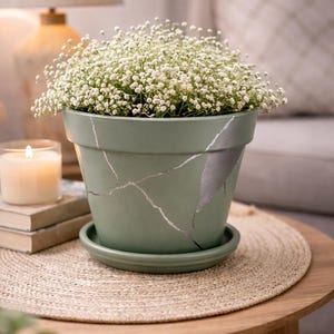 May include: A sage green ceramic plant pot with silver crackle detailing, filled with white baby's breath. The pot sits on a matching saucer, placed on a round, woven mat. A lit candle and books are in the background.