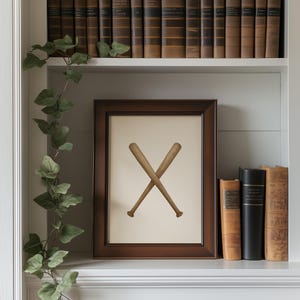 May include: A framed print featuring two crossed baseball bats in a dark brown wooden frame. The artwork is displayed on a white shelf, with a trailing green vine and a collection of antique books in the background.