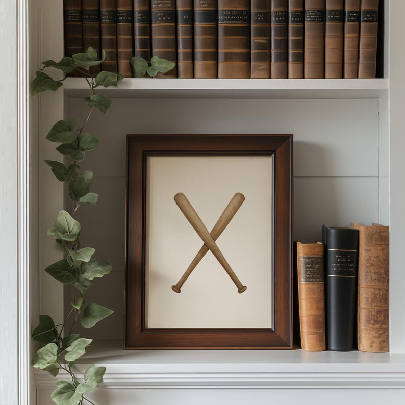 May include: A framed print featuring two crossed baseball bats in a dark brown wooden frame. The artwork is displayed on a white shelf, with a trailing green vine and a collection of antique books in the background.