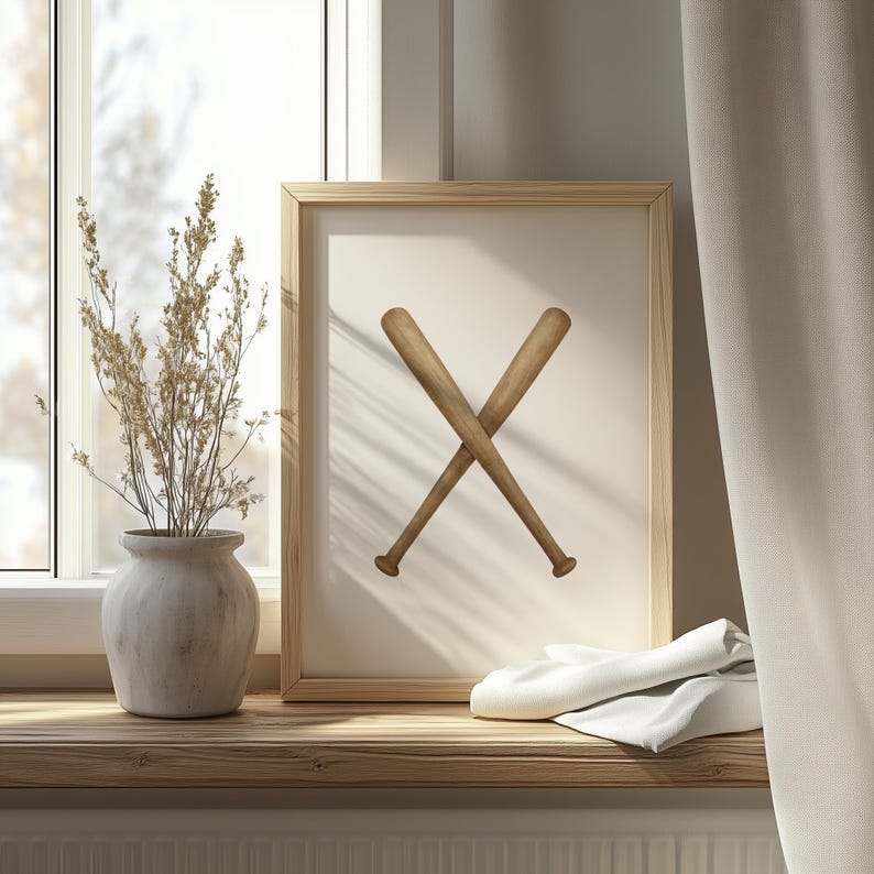 May include: A framed print featuring two crossed wooden baseball bats. The bats are light brown with darker brown accents, set against a cream-colored background. The frame is light wood. The print is on a wooden shelf with a vase and a white cloth.