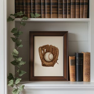 May include: A framed print featuring a baseball glove and ball. The artwork is in a brown wooden frame and rests on a white shelf. Decorative books and greenery are also visible, creating a classic, vintage aesthetic.
