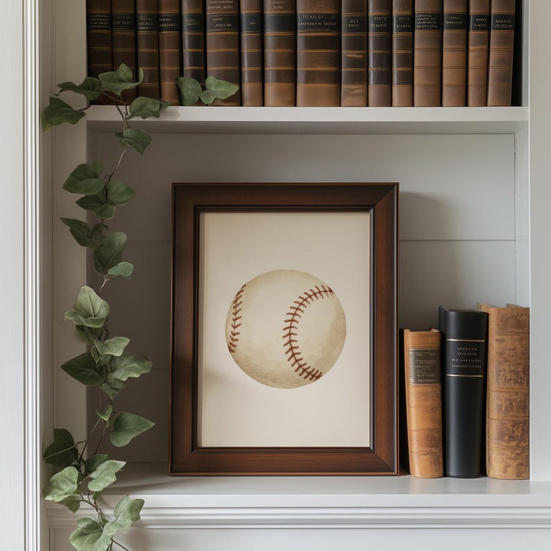 May include: A framed print of a baseball with red stitching. The print is in a dark brown wooden frame and sits on a white shelf with books and a trailing green vine. The baseball is off-white.