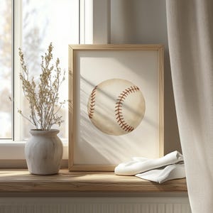 May include: A framed baseball print. The baseball is off-white with red stitching. The frame is light brown. The print is displayed on a wooden surface with a vase of dried flowers and a white cloth. The background includes a window and a curtain.