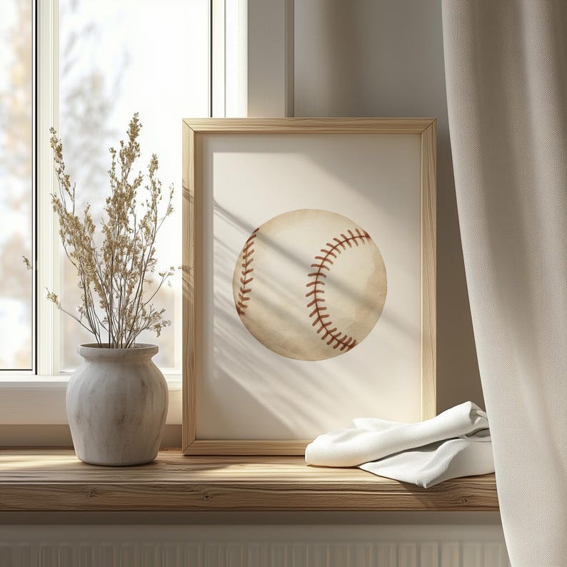 May include: A framed baseball print. The baseball is off-white with red stitching. The frame is light brown. The print is displayed on a wooden surface with a vase of dried flowers and a white cloth. The background includes a window and a curtain.