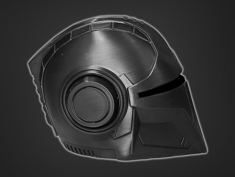 May include: A black helmet with a textured surface and a circular detail on the side. The helmet has a visor and a pointed chin guard.