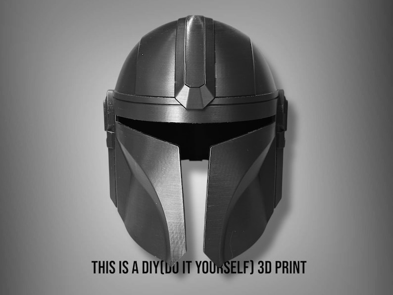 Puede incluir: Un casco mandaloriano negro y gris, una impresi&oacute;n 3D DIY. El casco tiene una visera en forma de T y rasgos angulares. El texto "THIS IS A DIY(DO IT YOURSELF) 3D PRINT" est&aacute; en la parte inferior.