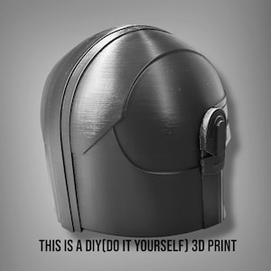 Puede incluir: Un casco negro y gris impreso en 3D con una superficie texturizada. El casco tiene una parte superior redondeada y un detalle tipo visera en el lateral. El texto en la parte inferior dice "THIS IS A DIY(DO IT YOURSELF) 3D PRINT."