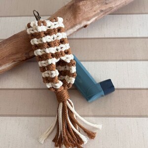 以下が含まれることがあります： 茶色と白の毛糸で編まれたタッセル付きのキーホルダー。キーホルダーは吸入器を入れるように設計されています。