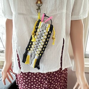 May include: A black, white, and yellow macrame glasses case with tassels hanging from the bottom. The case is attached to a lanyard with a gold-colored key chain.