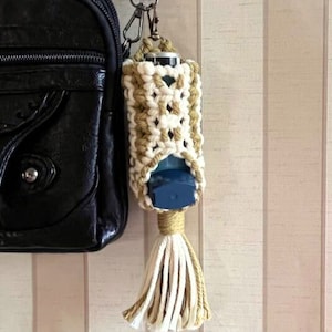 以下が含まれることがあります： 黒いバッグに付けられた、ベージュと白のタッセル付きのクロシェ編みのポーチに、吸入器が入っています。
