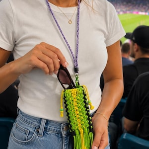 May include: A macrame sunglasses holder in black, green, and yellow, with a purple lanyard. The holder is rectangular with a tassel at the bottom and a drawstring closure. The lanyard is made of a braided cord.