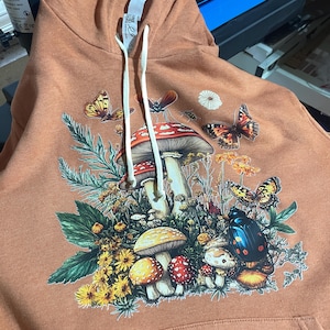 Könnte beinhalten: Rostfarbener Kapuzenpullover mit einem detaillierten Aufdruck von Pilzen, Schmetterlingen und Insekten. Das Design umfasst verschiedene Pilzarten, gelbe Blumen und Insektenillustrationen. Der Hoodie hat eine Kapuze mit Kordelzug und eine Vordertasche.