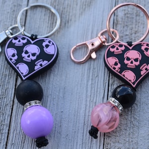 May include: Two keychains with black heart-shaped charms featuring pink skull designs. One keychain has a purple bead and the other has a pink bead.