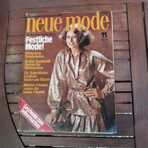 Può includere: Una copertina di rivista vintage con il titolo "neue mode" e il numero 11. La copertina presenta una donna in un abito fantasia. Il testo include "Festliche Mode!" e altro testo in tedesco. La rivista è su una superficie di legno.